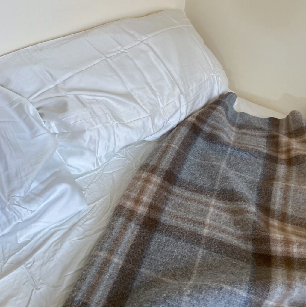 Two white pillows lay on white sheets, with a wool tartan blanket next to them. 